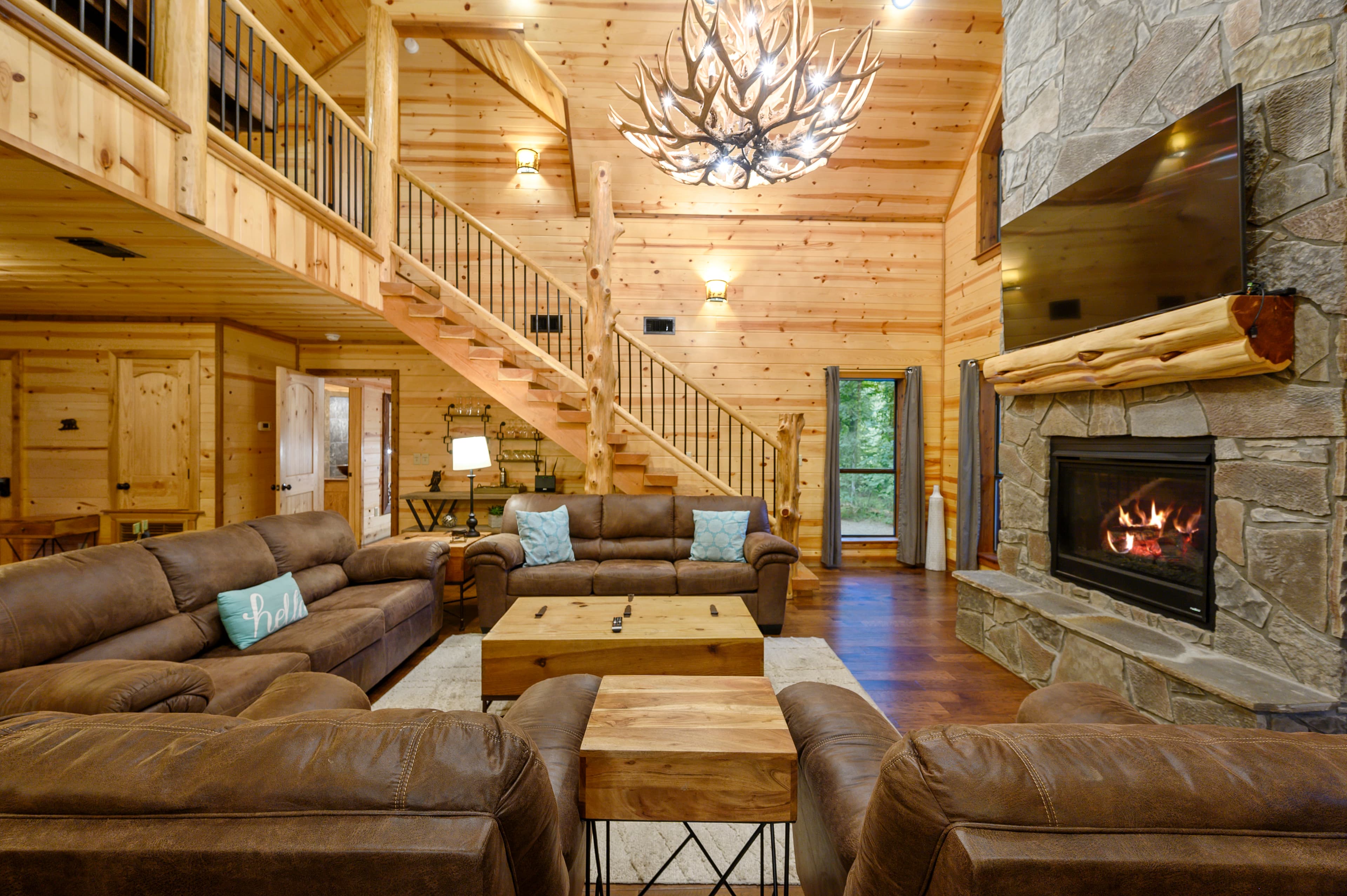 A cozy wooden cabin living room featuring leather sofas, a stone fireplace, and a large chandelier.