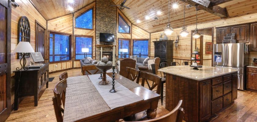 Open kitchen and dining area in the Beavers Bend Adventures cabin, Patio in the Pines, in Broken Bow, with a granite island and a stone fireplace under a vaulted wood ceiling.