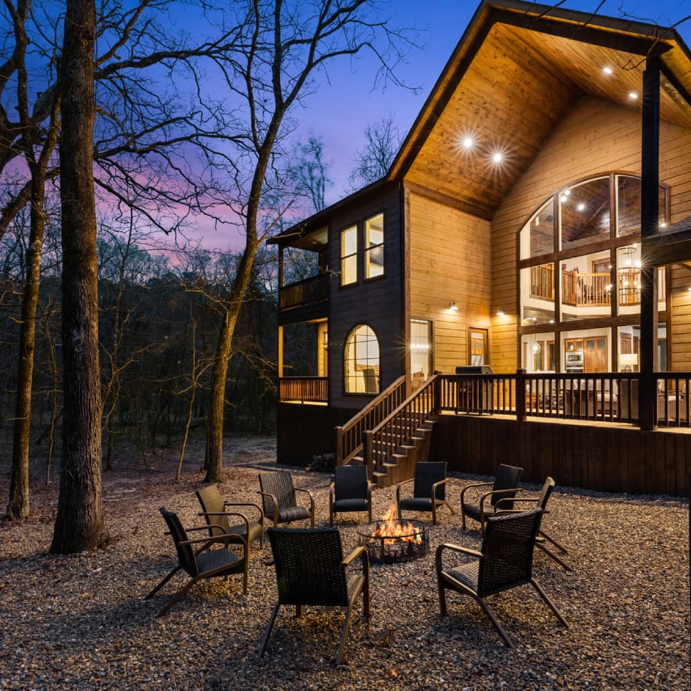 A cozy wooden house with large windows is illuminated at dusk, surrounded by trees and a fire pit with chairs.