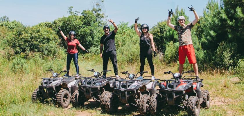 Four people in helmets stand atop ATVs in a lush green outdoor setting, smiling and posing with hand gestures.