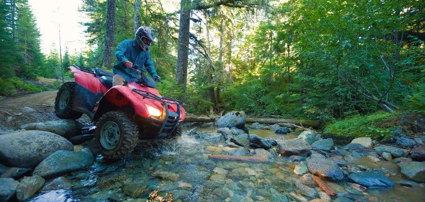 A person riding an ATV crosses a rocky stream in a forested area.