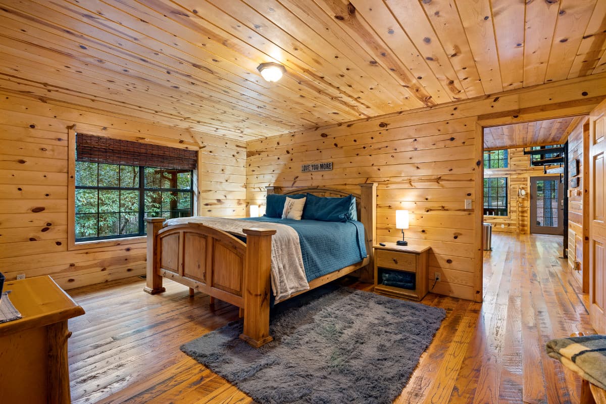 Cozy wooden bedroom featuring a large bed, plush rug, and natural light from windows.
