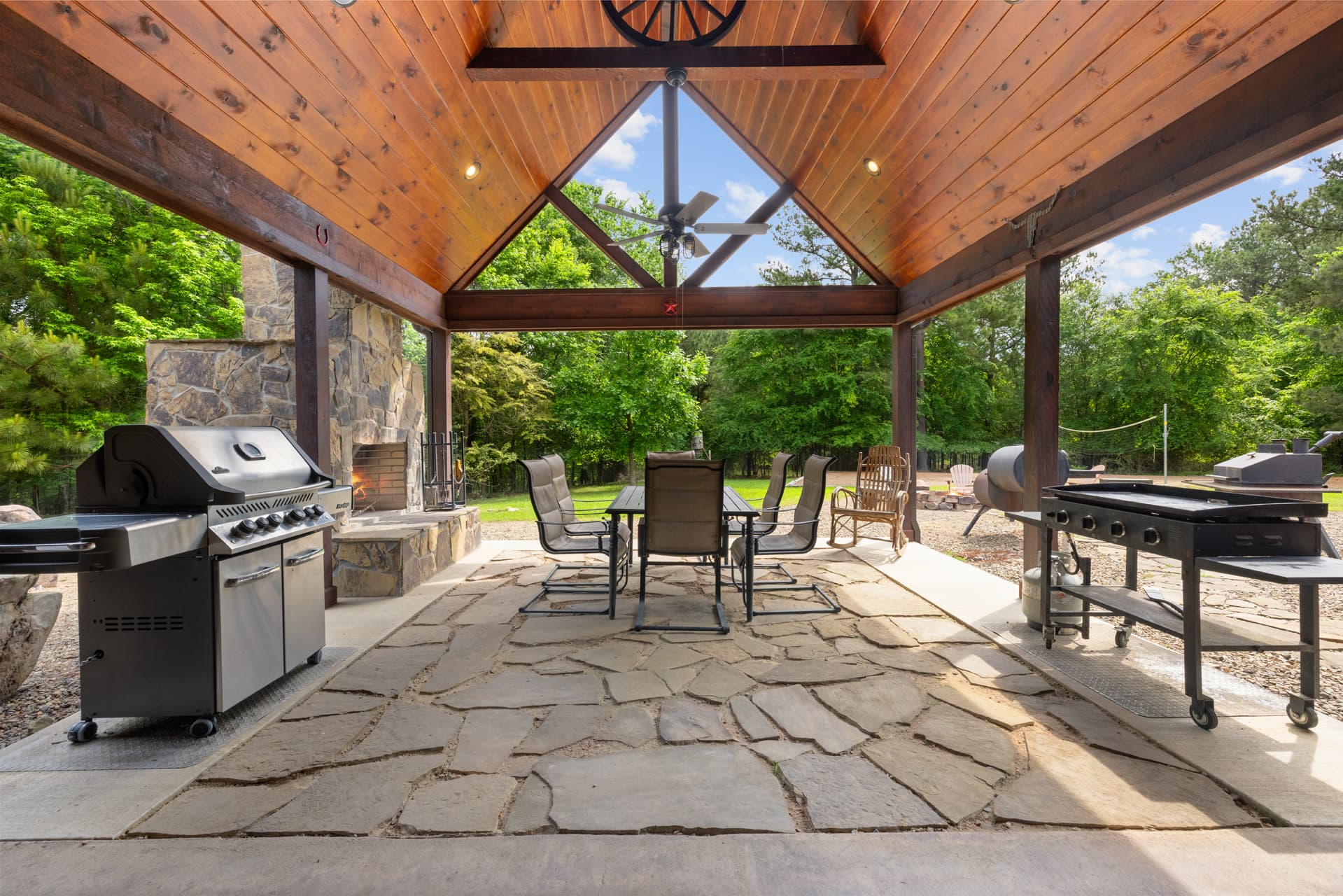 Outdoor dining area under a wooden pavilion with grills and a stone fireplace.