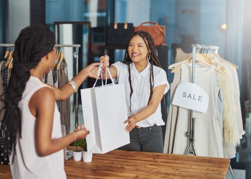 A smiling shopkeeper hands a shopping bag to a customer in a clothing store with a "SALE" sign. A smiling shopkeeper hands a shopping bag to a customer in a clothing store with a "SALE" sign.