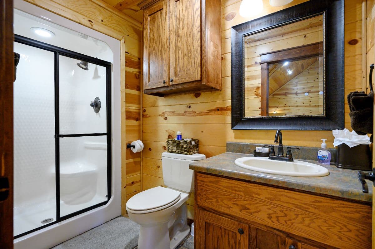 A rustic bathroom featuring wooden walls, a marble countertop, a shower with glass doors, and a mirror above the sink.