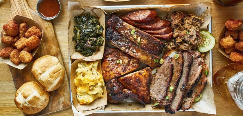 A barbecue tray piled with brisket, ribs, sausage, pulled pork, and smoked chicken, served with collard greens, mac and cheese, rolls, and fried bites.