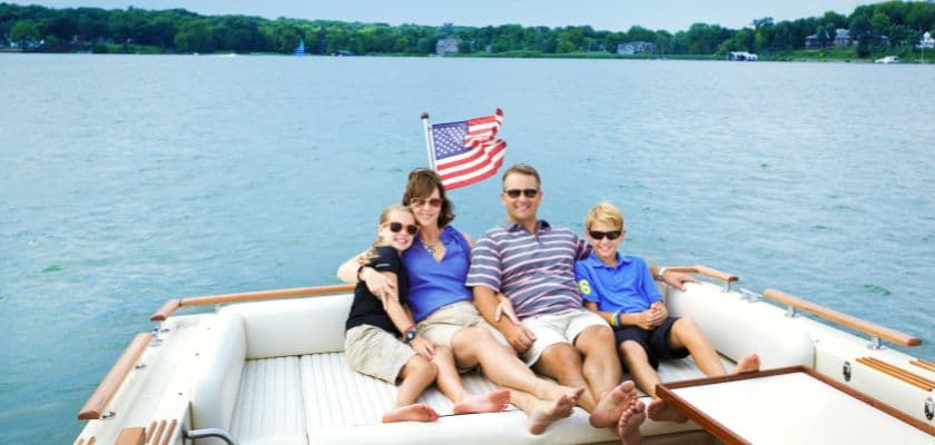 A family of four relaxes on a boat on a lake, with an American flag waving in the background.