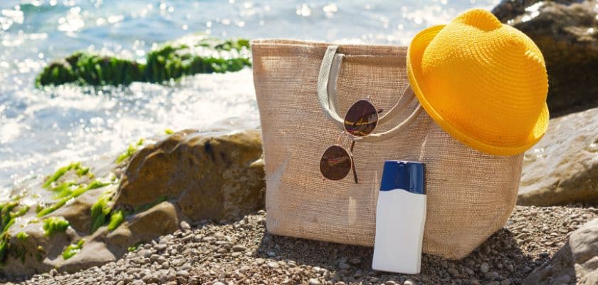 A beach scene featuring a straw hat, sunglasses, and sunscreen beside a tote bag on rocky shore.