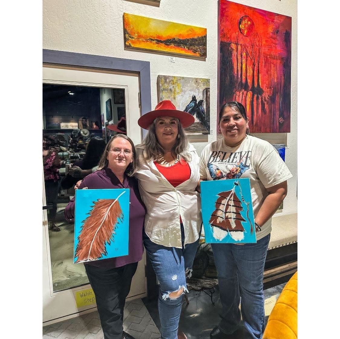 Three women holding paintings of feathers, standing in front of colorful artwork in a gallery.
