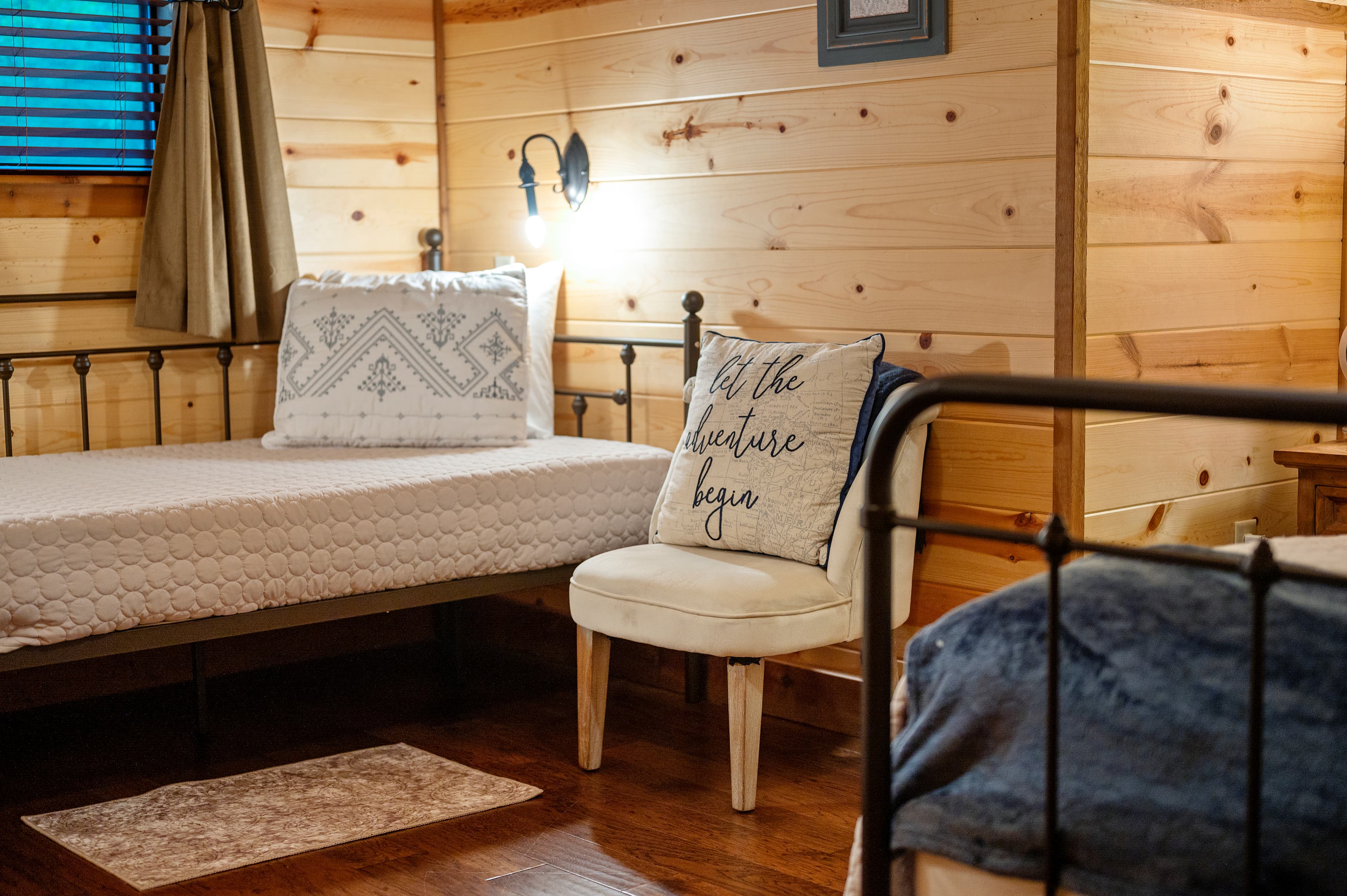 Cozy bedroom with two beds, a decorative pillow that says "Let the adventure begin," and wooden paneling.