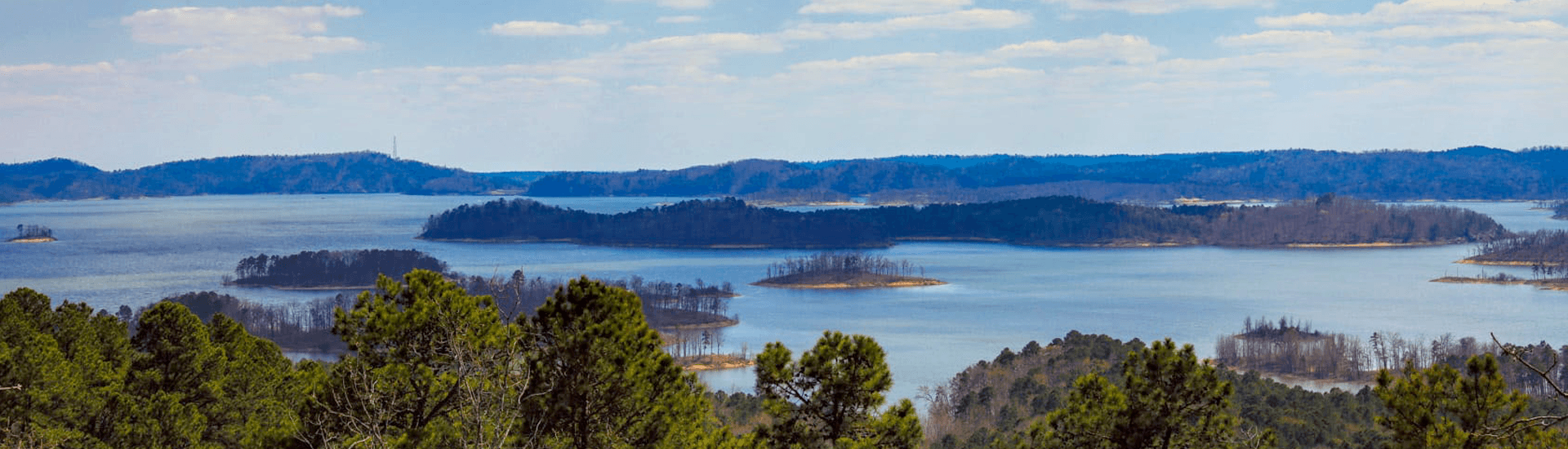 A panoramic view of a serene lake surrounded by forested islands and distant hills under a clear sky.