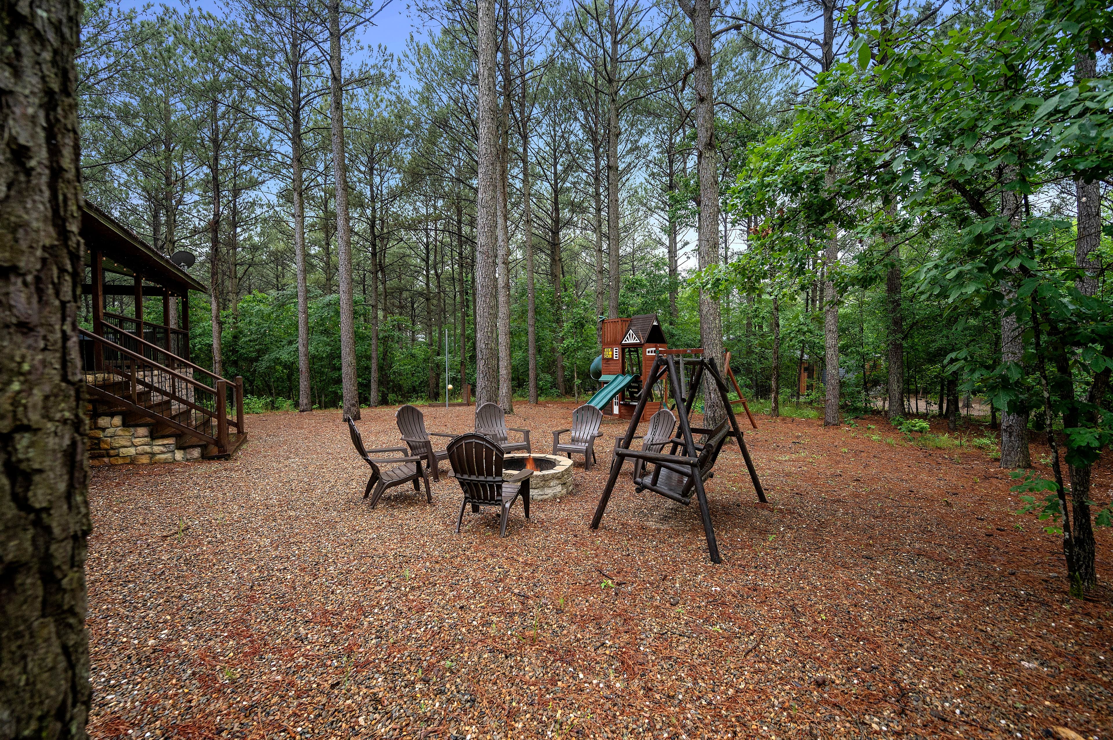 A seating area with chairs around a fire pit and a playground set in a wooded backyard.