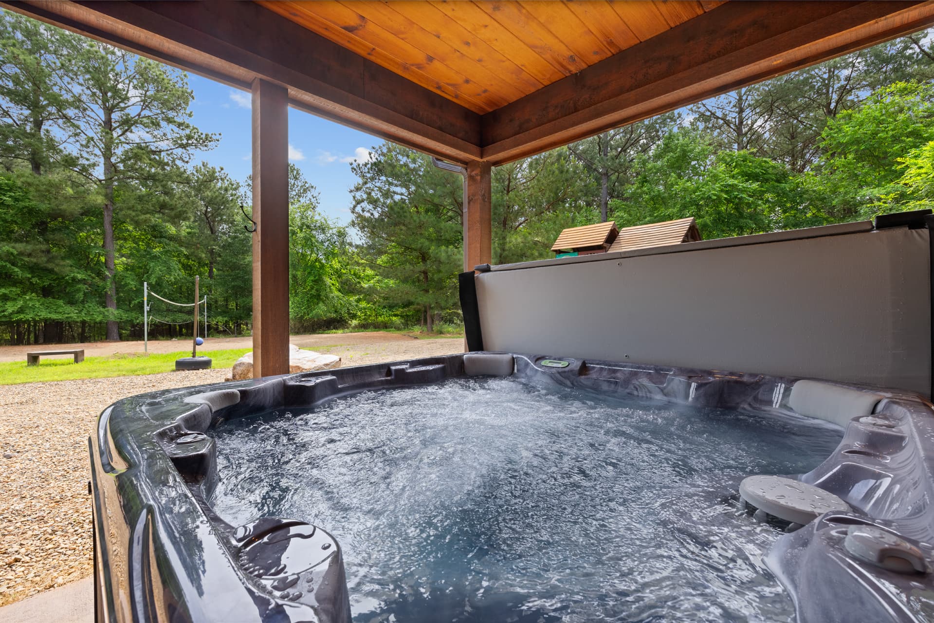 A hot tub sits under a porch, surrounded by trees and a grassy area.