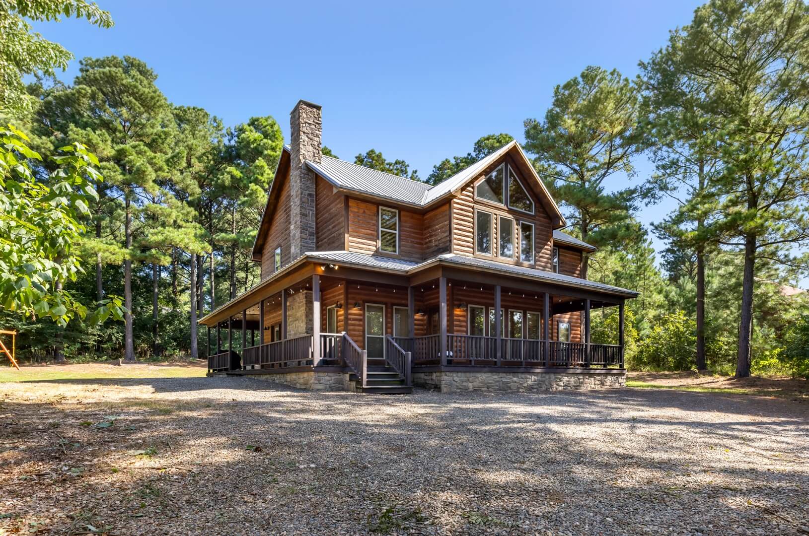 A large wooden house surrounded by tall trees and a gravel driveway. A large wooden house surrounded by tall trees and a gravel driveway.