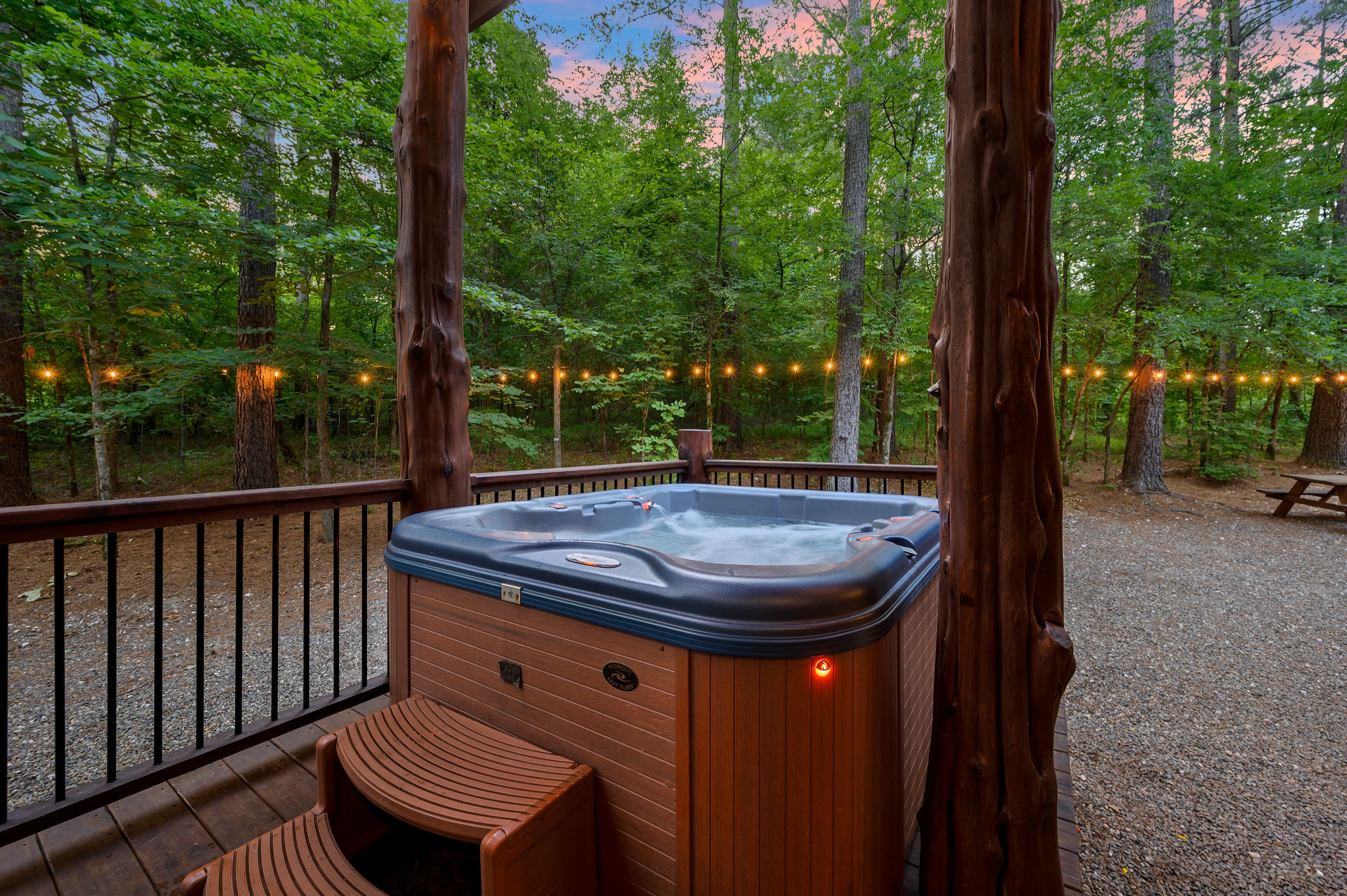 A hot tub on a wooden deck surrounded by trees, illuminated by string lights at dusk.