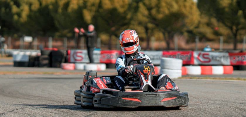 A young kart racer drives around a track, wearing a helmet and racing suit.