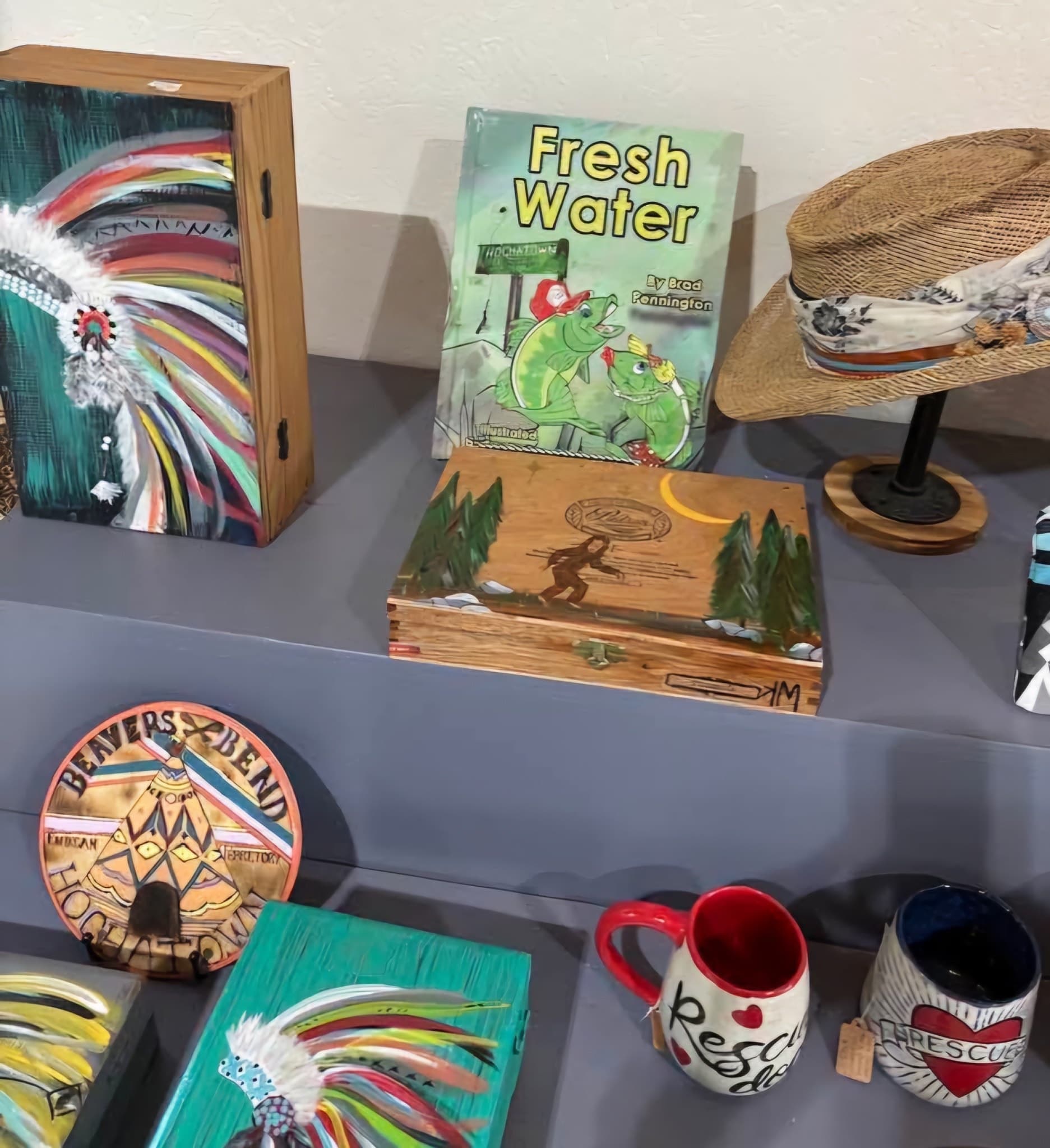 A display of various artworks, including painted boxes, mugs, and a children's book titled "Fresh Water."