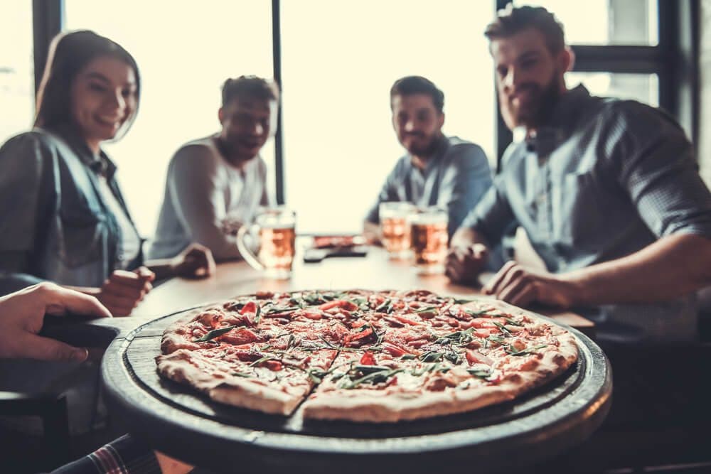 A freshly served pizza sits in the foreground while a group of friends enjoys their drinks in the background.