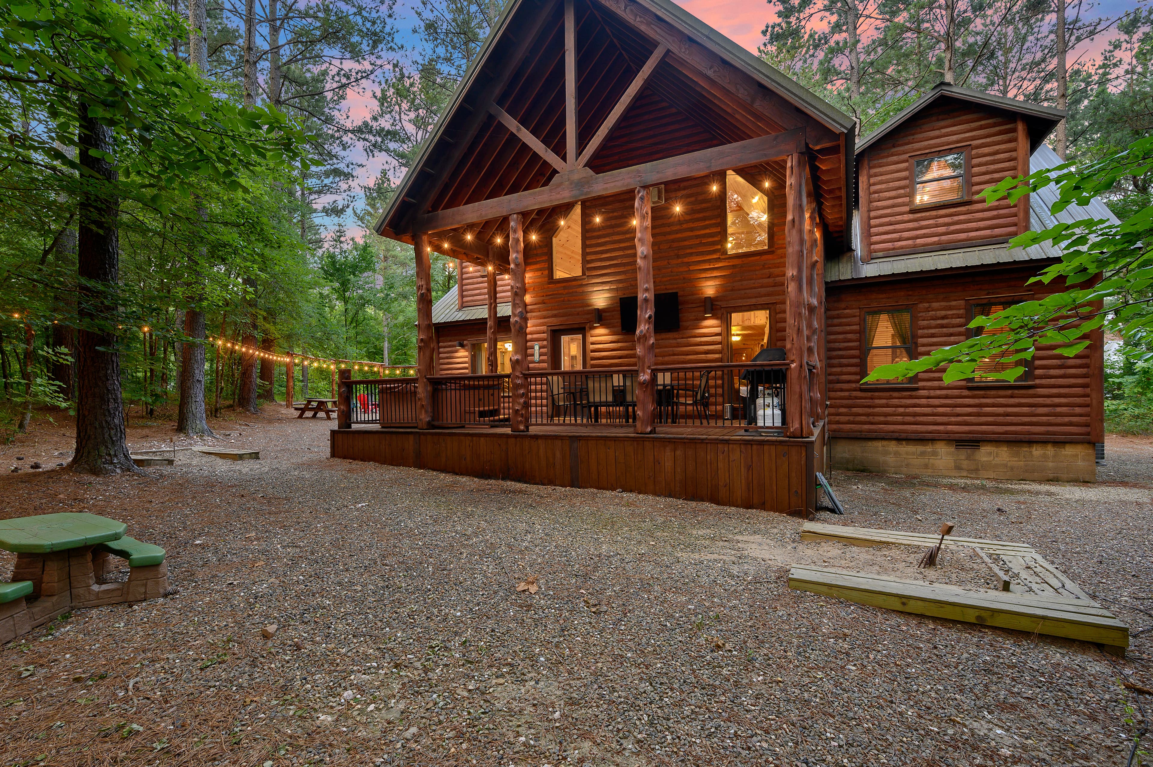 A large wooden cabin surrounded by trees, featuring a covered porch with string lights and a gravel yard.