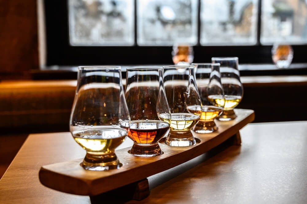 A wooden tasting flight holds five glasses of whiskey, arranged on a table with a backdrop of large windows.