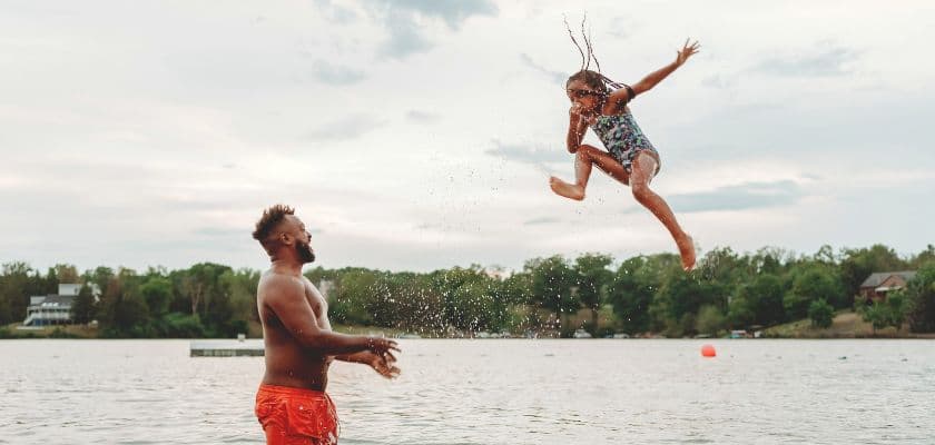 A girl joyfully leaps into a lake while a man stands below, ready to catch her.