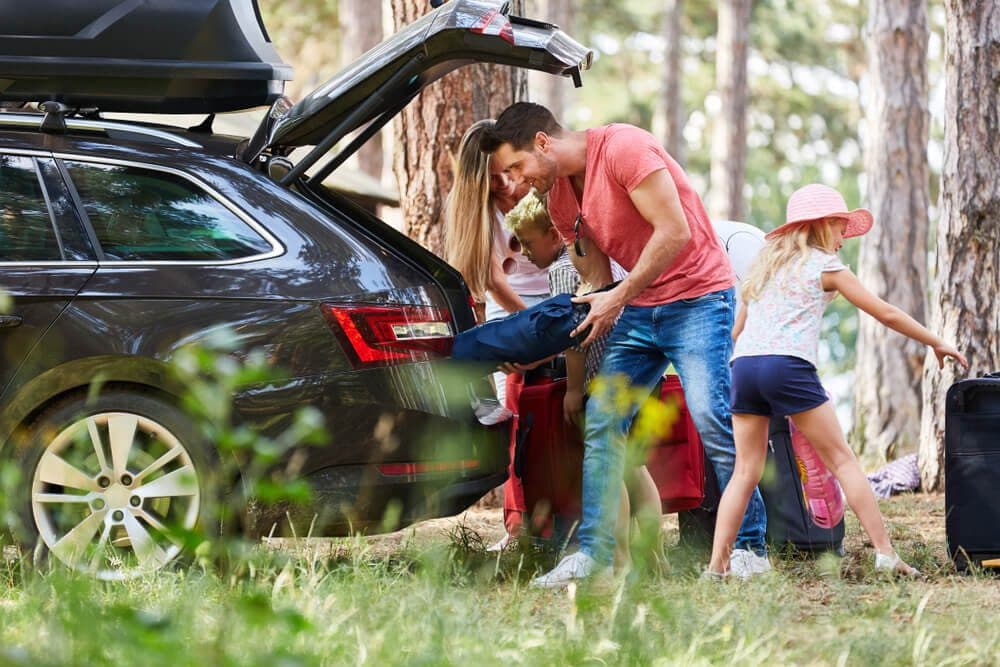 A family packs a car trunk for an outdoor trip in a forested setting.