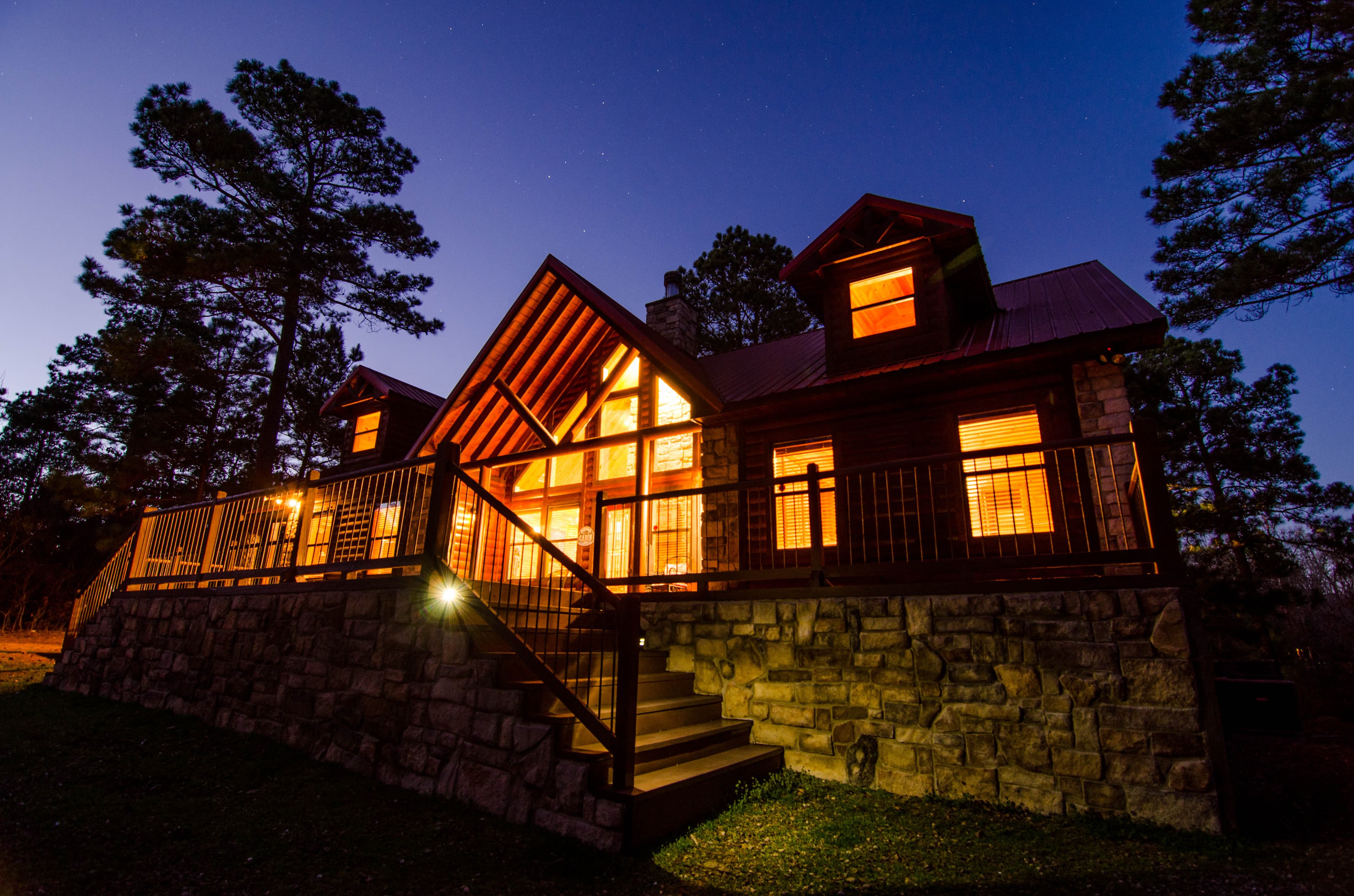 A warmly lit wooden cabin with a stone foundation, surrounded by trees at twilight.