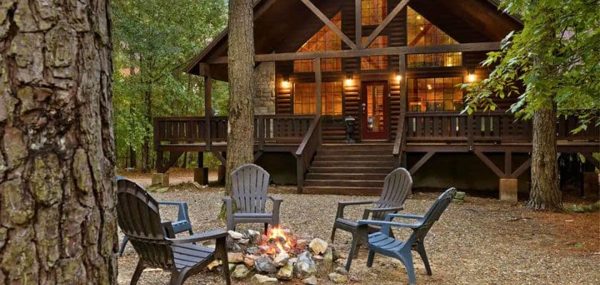 Log cabin surrounded by tall pine trees with Adirondack chairs gathered around a fire pit.