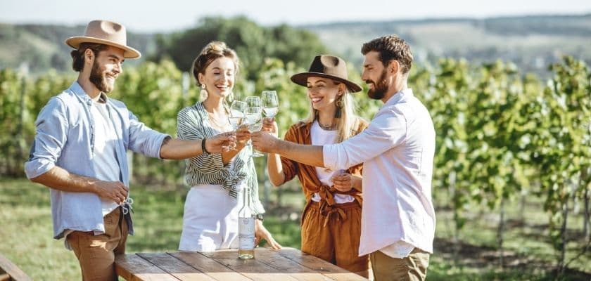 two couples standing in a vineyard, toasting with glasses of white wine
