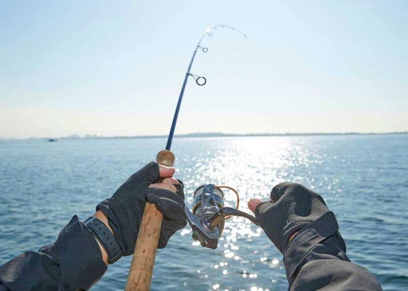 Hands wearing gloves hold a fishing rod and reel over the water, with sunlight reflecting across the lake's surface. Hands wearing gloves hold a fishing rod and reel over the water, with sunlight reflecting across the lake's surface.