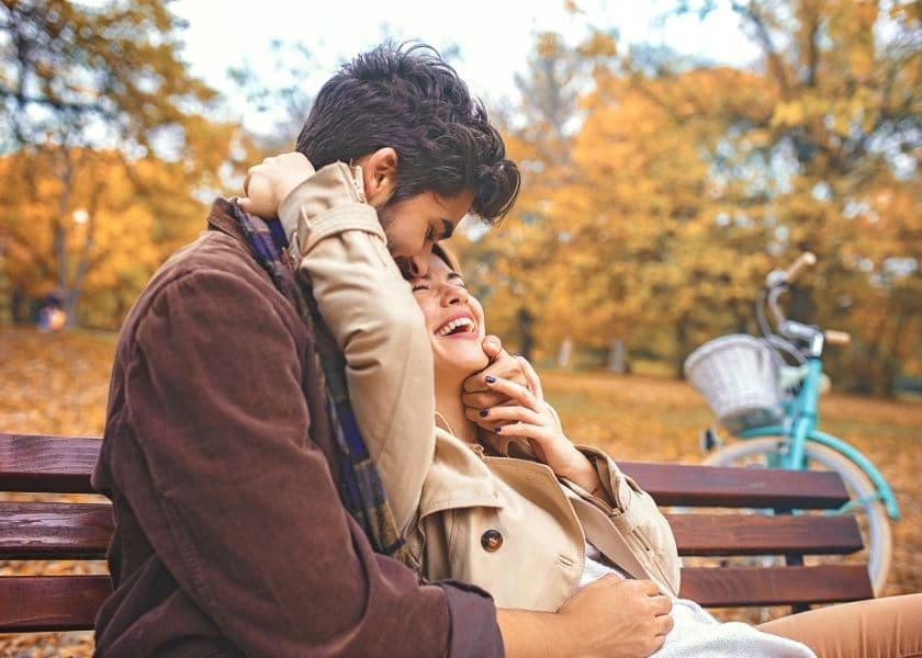A couple sitting on a park bench, embracing beneath colorful fall trees with a bicycle nearby.